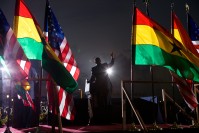 Barack Obama on stage, waving at the crowd, surrounded by American and Ghanaian flags.