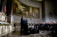Barack Obama speaking to an audience. In the background, a mural depicting the writing and adoption of the Declaration of Independence and the Constitution of the United States.
