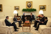 Barack Obama and Manmohan Singh, seating next to each other leg crossed, talking. Next to each of them, two men taking notes.