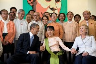Barack Obama, Hilary Clinton and Aung San Suu Kyi, holding hands, seating together with the staff standing behind them.