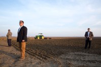 Three men wearing sunglasses, standing in a field. In the background, a tractor.